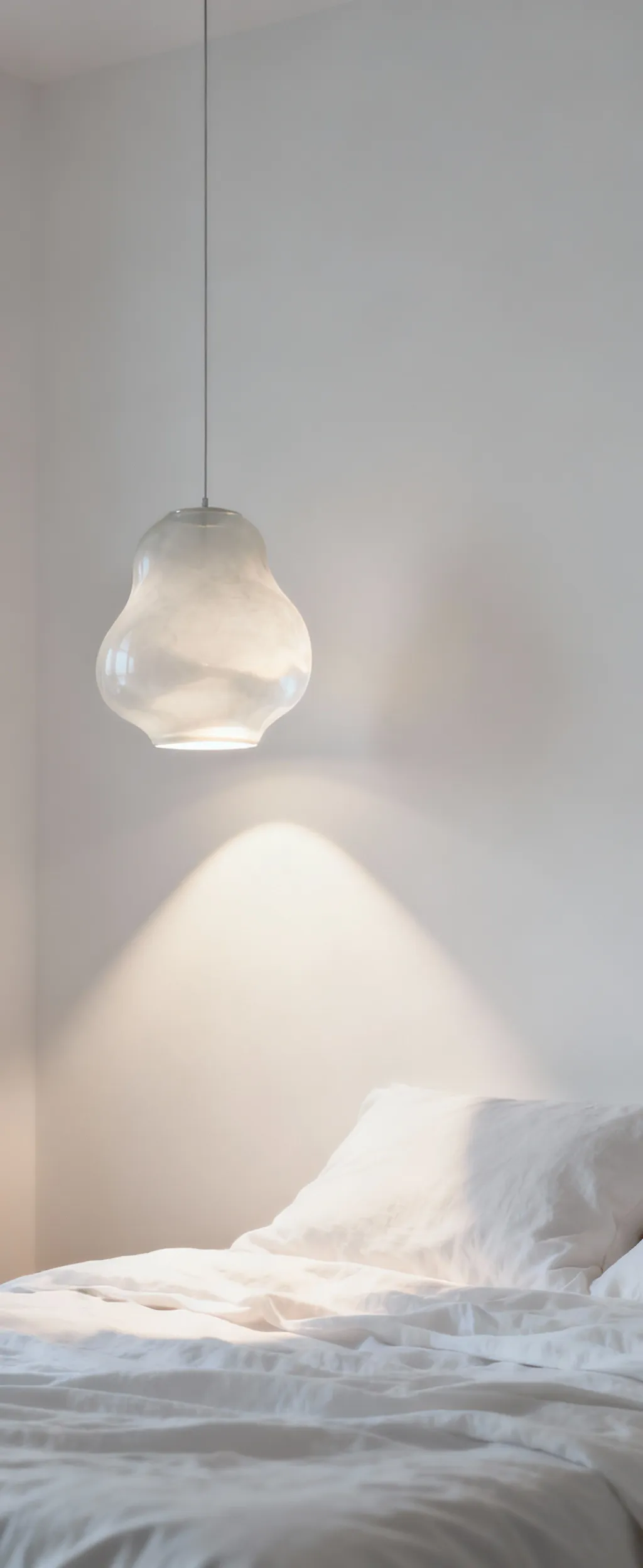 A serene white bedroom features a sculptural opaque glass pendant, illuminated by precise spot lighting that casts soft shadows on the white wall, creating depth and dimension.