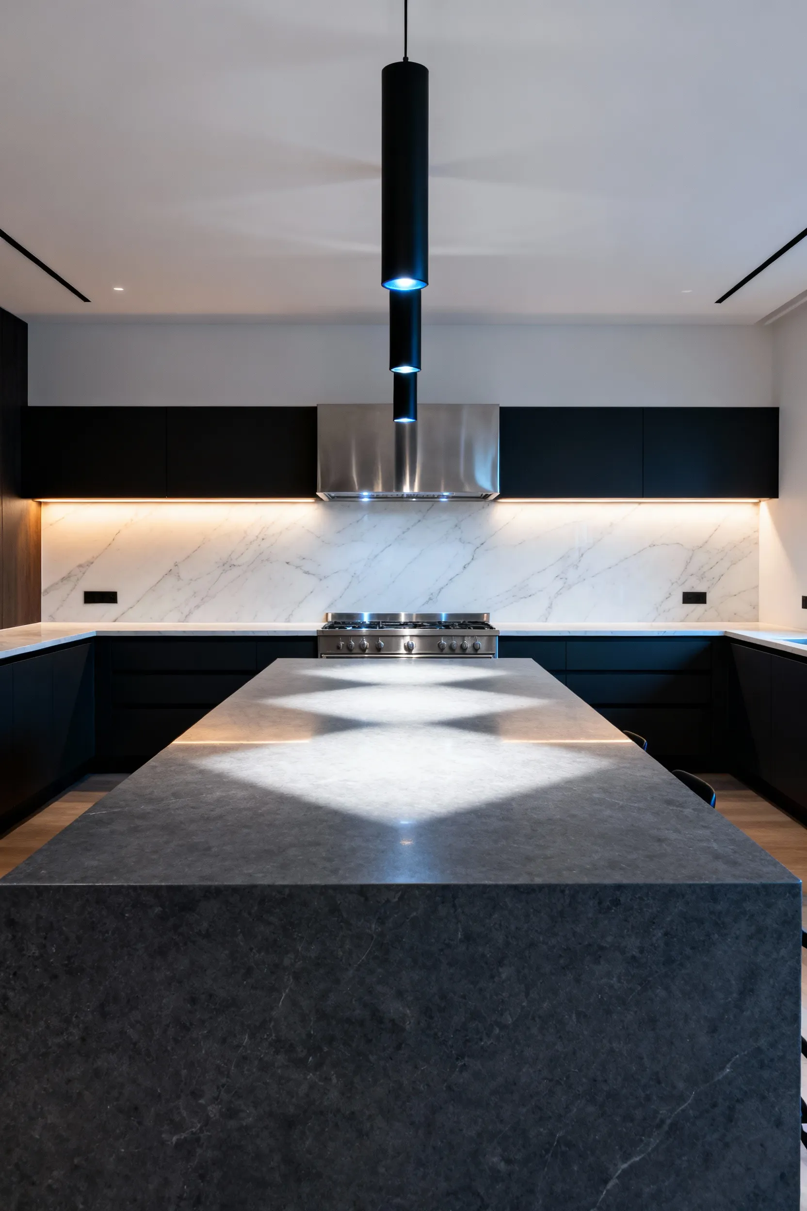 A wide view of a modern kitchen showing three black cylinder pendant lights centered directly beneath a dominant stainless steel range hood, balancing the sightline above a large gray kitchen island.