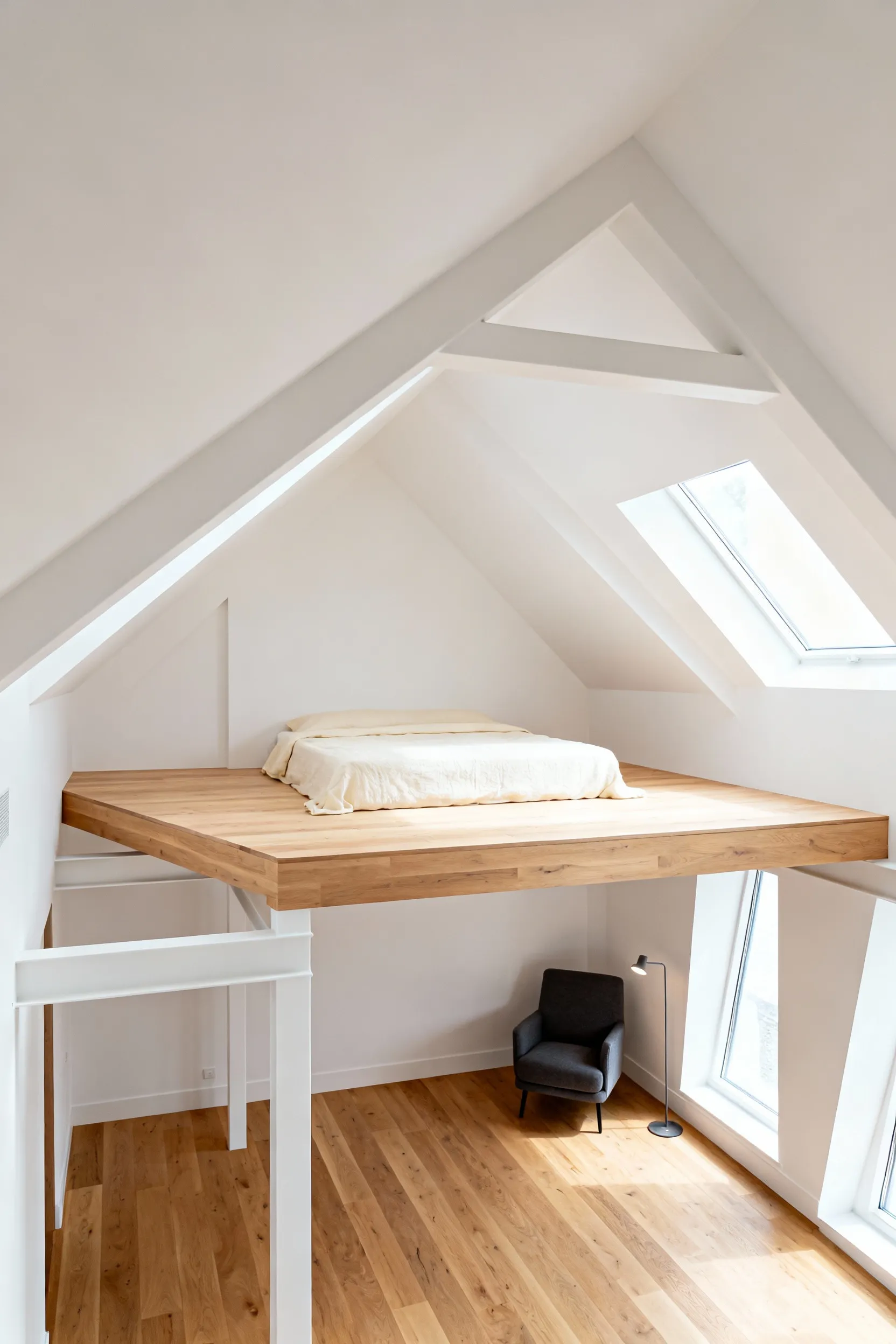 Elevated bleached oak platform bed in a modern white attic bedroom, illustrating multi-level zoning and integrated storage underneath the raised sleeping area.