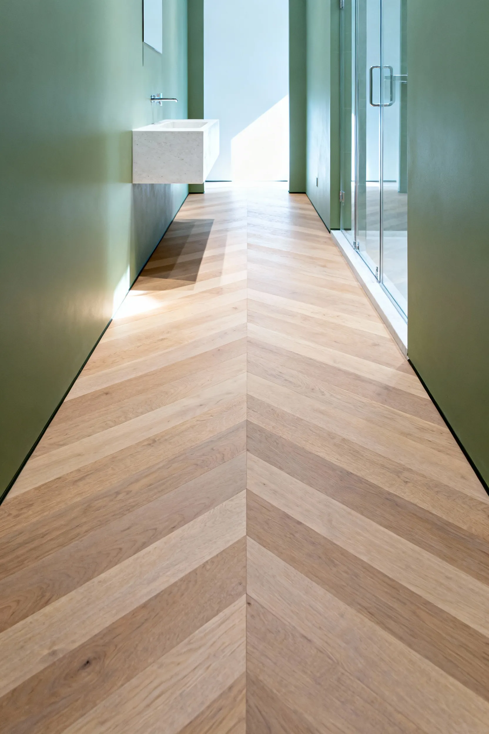 A long, narrow galley bathroom featuring light oak wood flooring installed in a directional chevron pattern that visibly elongates the space, leading the eye toward a bright back wall.