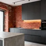 A modern kitchen interior featuring an exposed red brick accent wall that contrasts sharply with sleek stainless steel appliances and minimalist gray cabinetry.