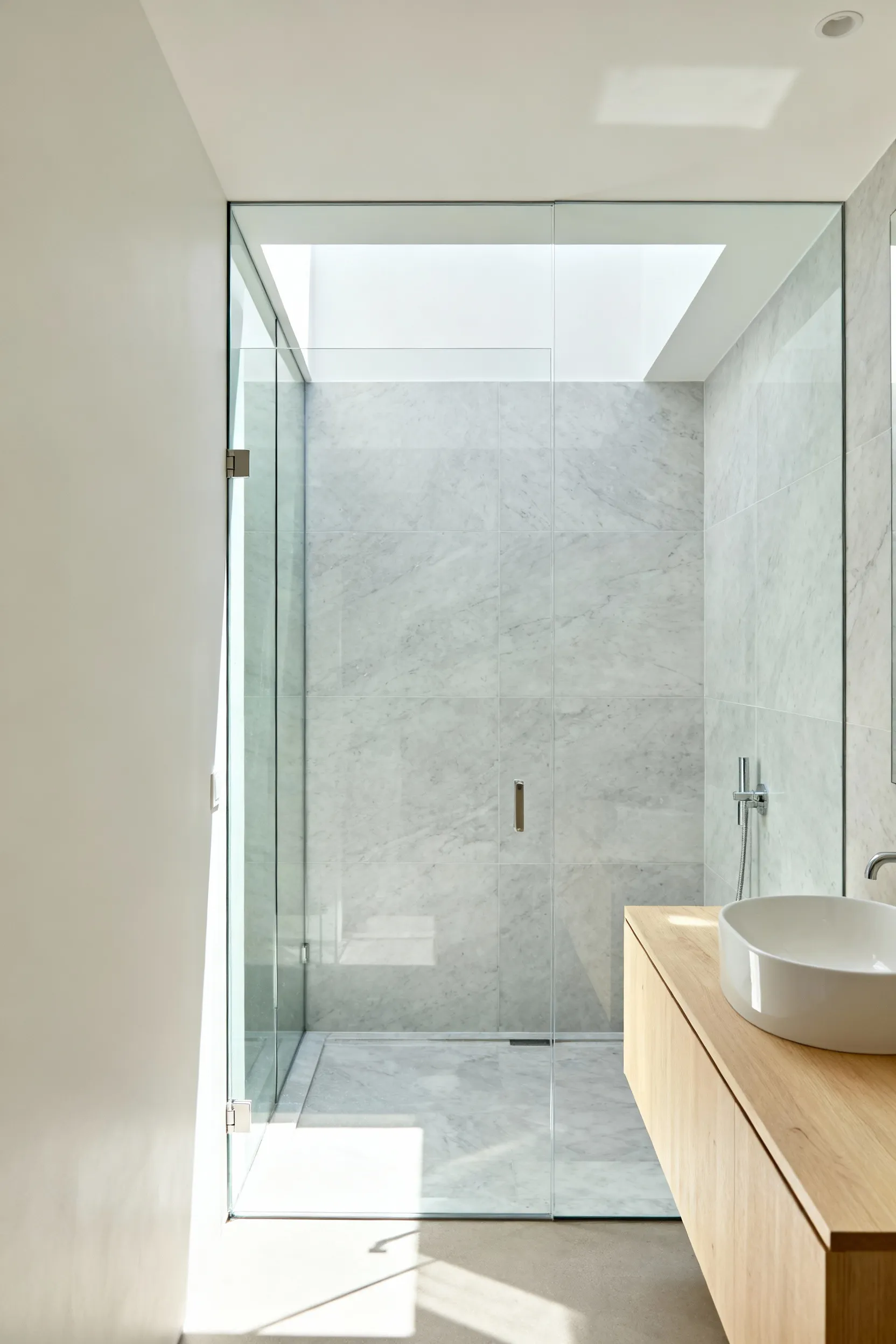 A small modern bathroom featuring a completely frameless, transparent glass shower partition designed to eliminate visual barriers and extend sightlines, creating an illusion of a much larger room.
