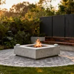 A beautifully integrated backyard firepit area, strategically placed for optimal ambiance and safety, featuring natural stone seating and lush landscaping, under soft evening light.