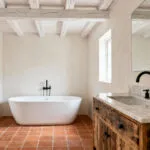 A sophisticated farmhouse bathroom featuring a reclaimed wood vanity with honed marble and matte black fixtures, alongside a white freestanding tub on terracotta-look porcelain tiles, showcasing rustic and contemporary design integration.