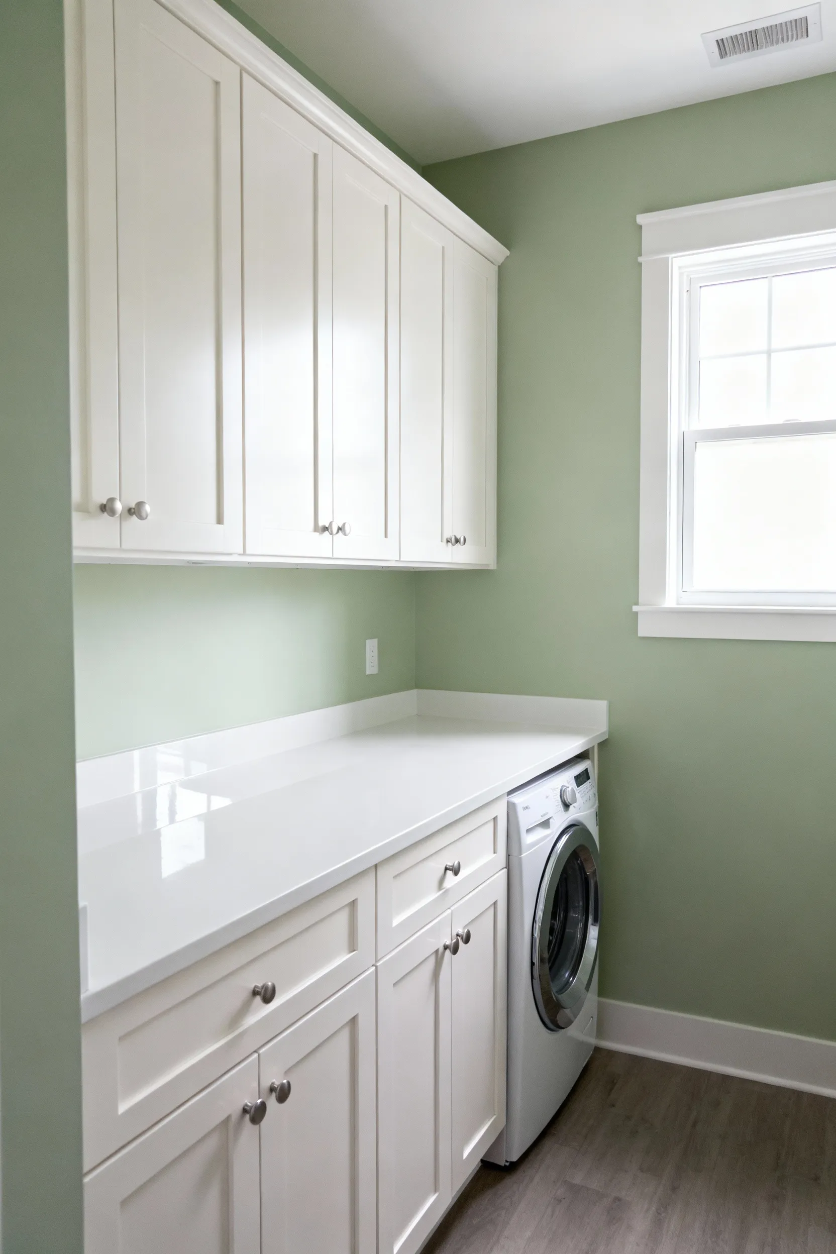A serene, compact laundry room featuring soft sage green walls, pure white custom cabinetry, and a reflective white countertop, embodying a calming color palette for enhanced spatial comfort.
