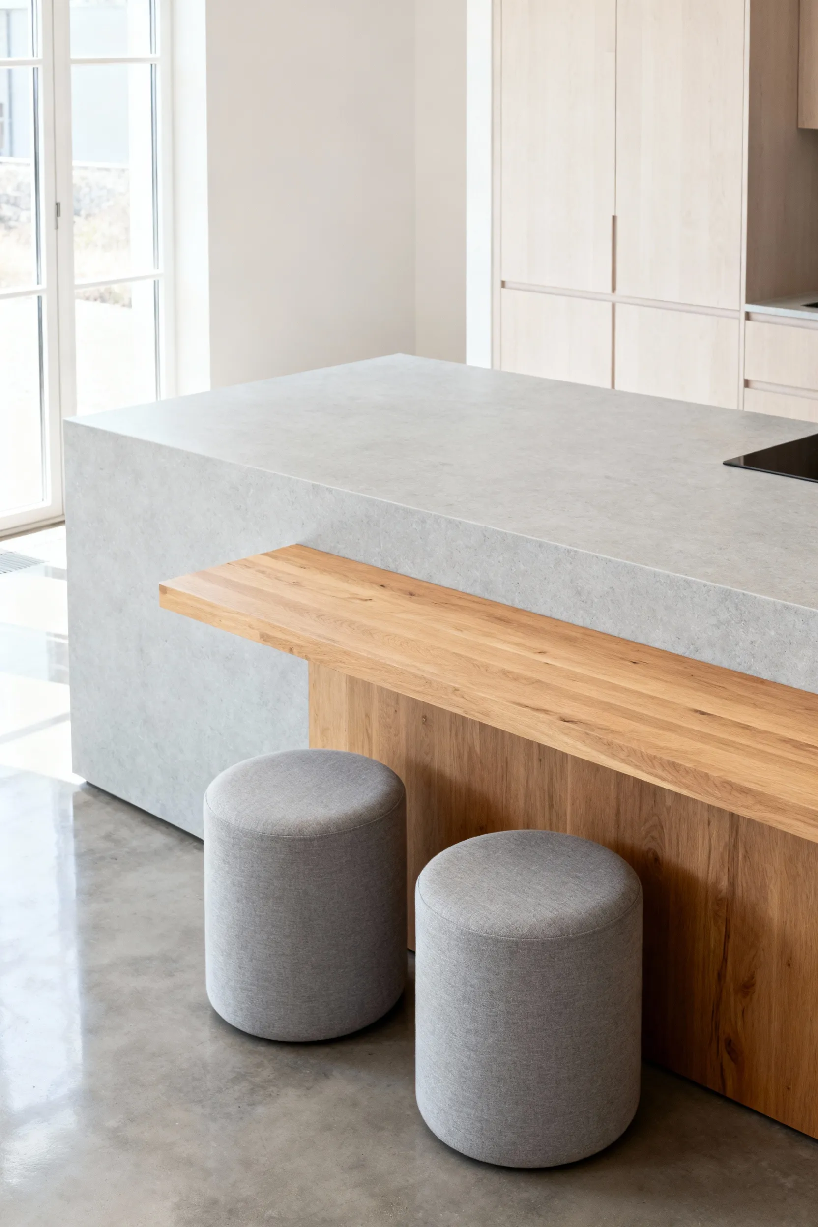 A bright Scandinavian kitchen with a large light grey honed quartz island featuring an integrated solid ash wood extension, accompanied by two grey linen upholstered stools.