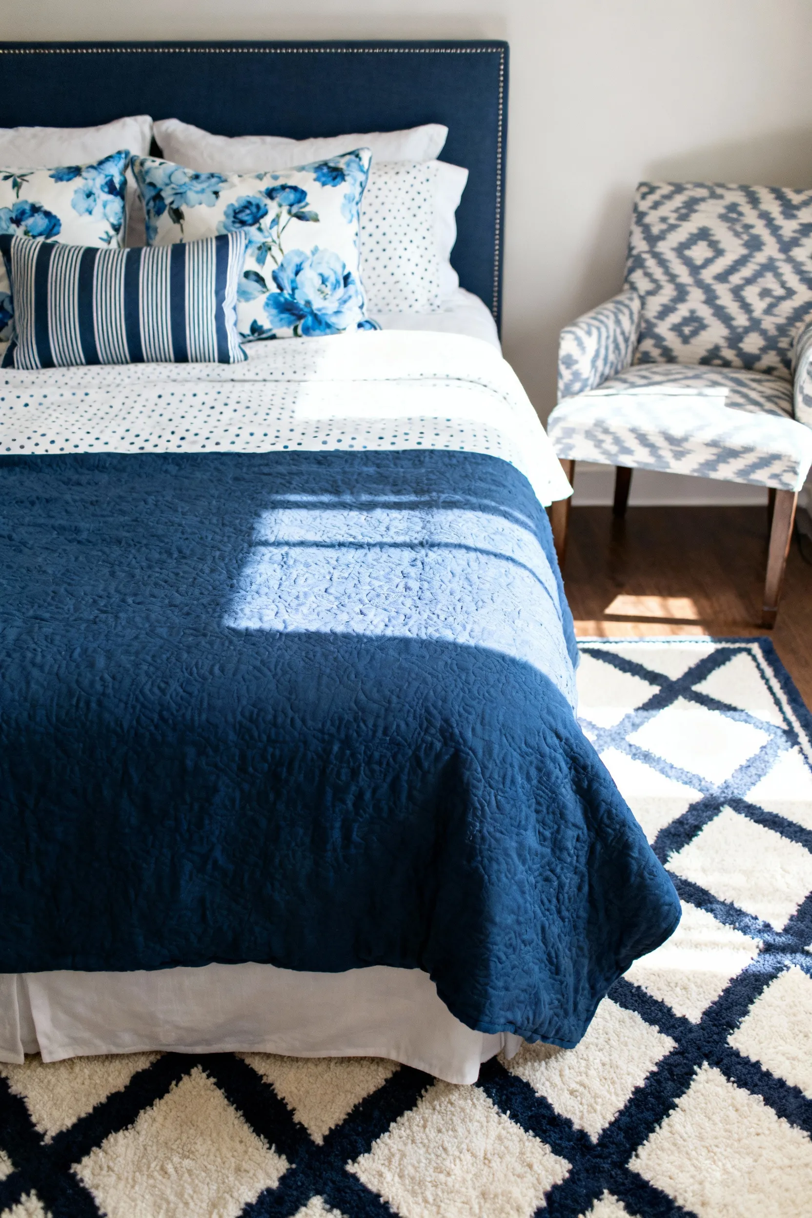 A sophisticated blue bedroom featuring strategic pattern mixing, including a large geometric rug, small-scale floral throw pillows, and an ikat accent chair, all maintaining a cohesive blue and white color palette.