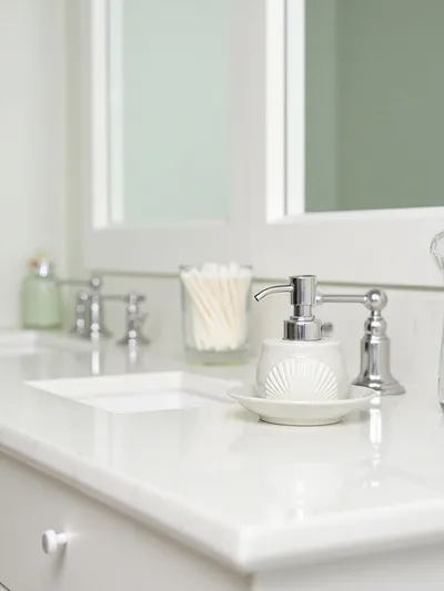 Close-up of a clean coastal bathroom vanity countertop featuring a ceramic soap dispenser with a seashell design and a small glass jar with cotton swabs, illuminated by soft, even lighting.