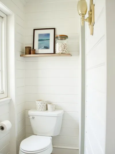Charming coastal bathroom featuring a shelf displaying framed beach photos and a glass jar of seashells, set against white shiplap walls and light wood accents, illuminated by natural daylight.