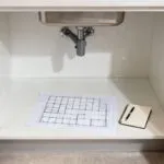 A meticulously clean, empty under-sink kitchen cabinet with a conceptual spatial grid drawing and a notebook, representing the crucial initial steps of a complete decluttering purge and inventory for kitchen organization.