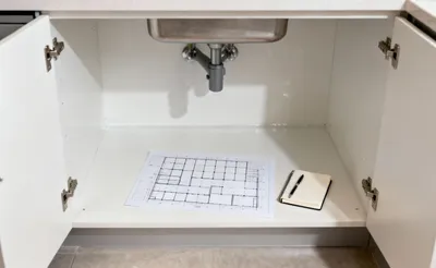A meticulously clean, empty under-sink kitchen cabinet with a conceptual spatial grid drawing and a notebook, representing the crucial initial steps of a complete decluttering purge and inventory for kitchen organization.