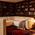A warm, intimate living room nook featuring built-in dark wood bookshelves and a cream sofa, designed to create a protected sense of refuge and psychological enclosure.
