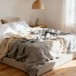 A rumpled bed layered with high-quality, textured natural linen sheets in grey and white tones, showcasing the durable and breathable foundation for a stylish wabi-sabi bedroom aesthetic.