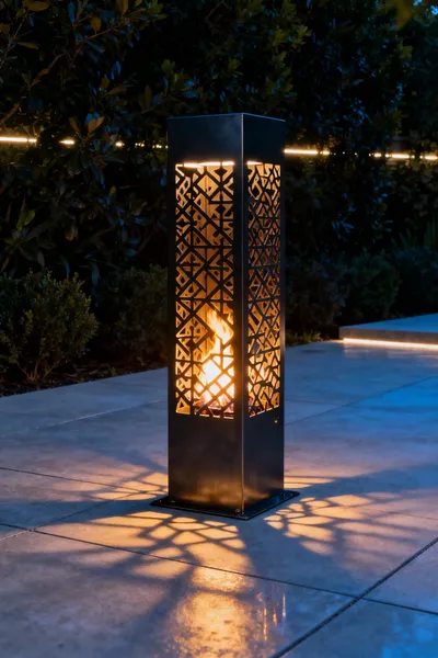 Modern outdoor firepit with a tall, anodized aluminum perforated metal enclosure, casting dramatic shadows on concrete paving at twilight from the flickering fire and subtle LED backlighting.