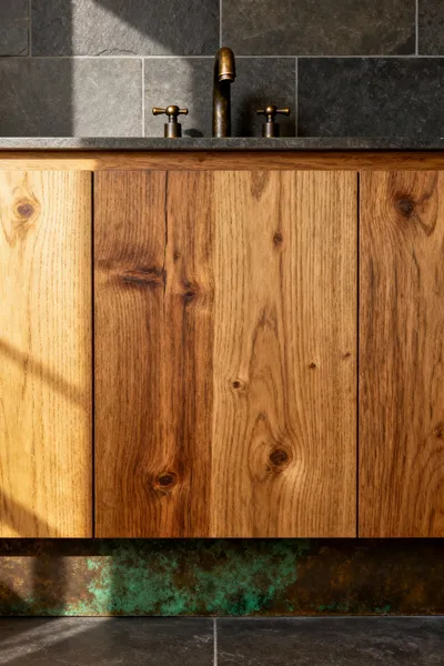 Farmhouse bathroom vanity with unlacquered brass faucets displaying a natural patina and a custom white oak vanity top against honed slate tiles, emphasizing aged character and authenticity.