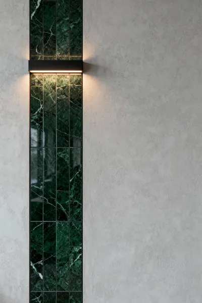 Luxury bathroom wall featuring a contrasting design with a polished dark green marble panel embedded into a matte light grey micro-cement wall, showcasing a curated disruption aesthetic. Modern architectural lighting subtly illuminates the textures and reflective differences.