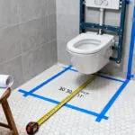 A photograph of a small, renovated bathroom with light gray tile showing measuring tape and blue lines on the floor indicating the precise 30-inch clearance zone required for a toilet fixture according to building codes.