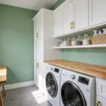 A brightly lit modern laundry room featuring an ergonomic layout with white cabinets, a wood folding counter, and organized storage at reaching height.